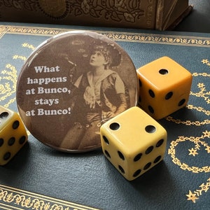 May include: A brown button with the text "What happens at Bunco, stays at Bunco!" and a vintage photo of a woman in a cowboy hat. There are three yellow dice on a dark blue surface.
