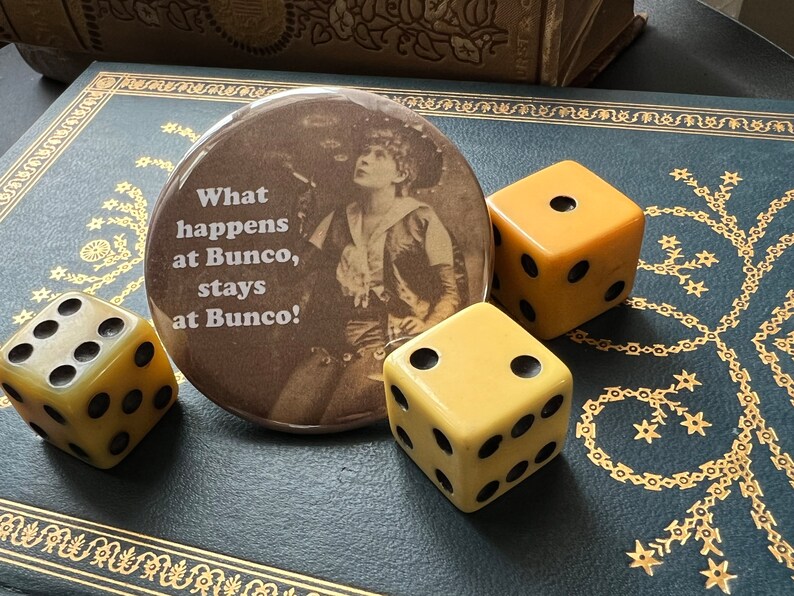May include: A brown button with the text "What happens at Bunco, stays at Bunco!" and a vintage photo of a woman in a cowboy hat. There are three yellow dice on a dark blue surface.