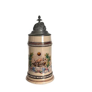 May include: A beige ceramic beer stein with a pewter lid. The stein features a colorful illustration of a pig, sun, and text. The top and bottom have decorative bands. The stein is approximately 10 inches tall.