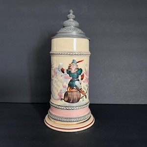 May include: A vintage beer stein with a hinged metal lid. The ceramic body is cream-colored with a painted scene of a man in a green and red outfit. The stein has a pink and cream base with decorative trim.