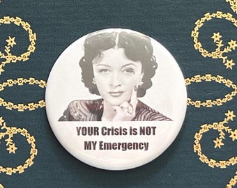 May include: A white button with a black and white image of a woman and the text "YOUR Crisis is NOT MY Emergency". The button is on a dark green surface with gold star-shaped embellishments.