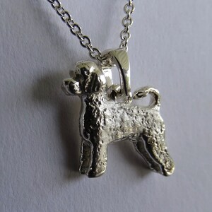 May include: Silver pendant necklace featuring a detailed, textured, standing dog charm. The dog is a breed with curly hair, and the pendant is attached to a silver chain.