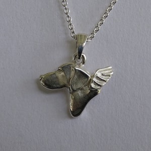 Może przedstawiać: Srebrny naszyjnik z zawieszką przedstawiającą głowę psa ze skrzydłem. Zawieszka jest przymocowana do delikatnego srebrnego łańcuszka. Głowa psa jest szczegółowa, a skrzydło dodaje fantazyjnego akcentu. Naszyjnik jest prawdopodobnie wykonany ze srebra próby 925.
