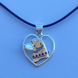 Puede incluir: Collar con colgante de plata en forma de corazón, que representa un perfil de cabeza de perro detallado con un collar de piedras preciosas rojas.