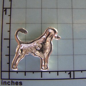Puede incluir: Broche de metal plateado de un perro, posiblemente un Bloodhound, de pie de perfil. La broche mide aproximadamente 2,5 cm de alto.