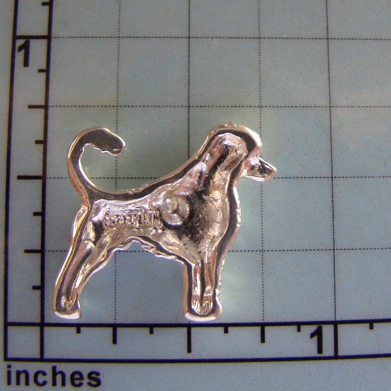 Puede incluir: Broche de metal plateado de un perro, posiblemente un Bloodhound, de pie de perfil. La broche mide aproximadamente 2,5 cm de alto.