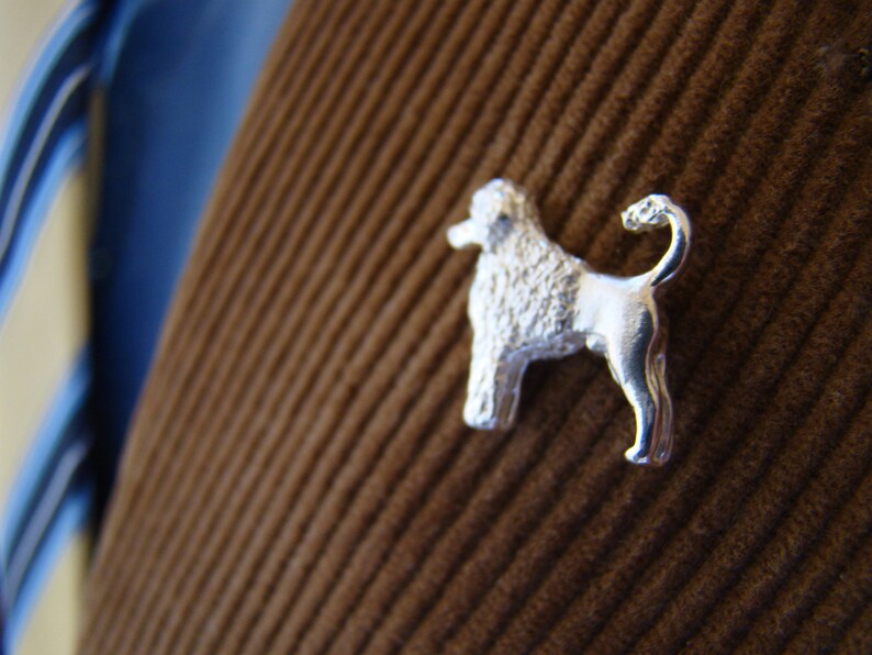 Puede incluir: Un broche de perro caniche de color plateado, con una cola rizada, est&aacute; fijado a una tela de pana marr&oacute;n.