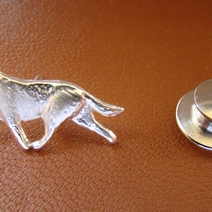 May include: A silver-colored pin featuring a detailed depiction of a German Shepherd dog in a running pose. The pin is accompanied by a separate, round, silver-colored backing piece. The items are displayed on a brown surface.