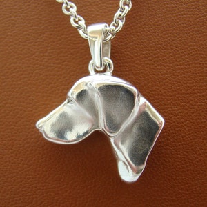 Può includere: Collana con ciondolo in argento con un designo dettagliato di testa di cane in tre dimensioni. Il ciondolo è attaccato a una catena d'argento.