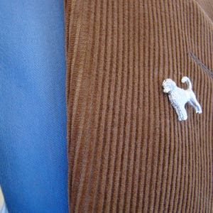 Puede incluir: Una chaqueta de pana marr&oacute;n con un broche de perro plateado en la solapa.