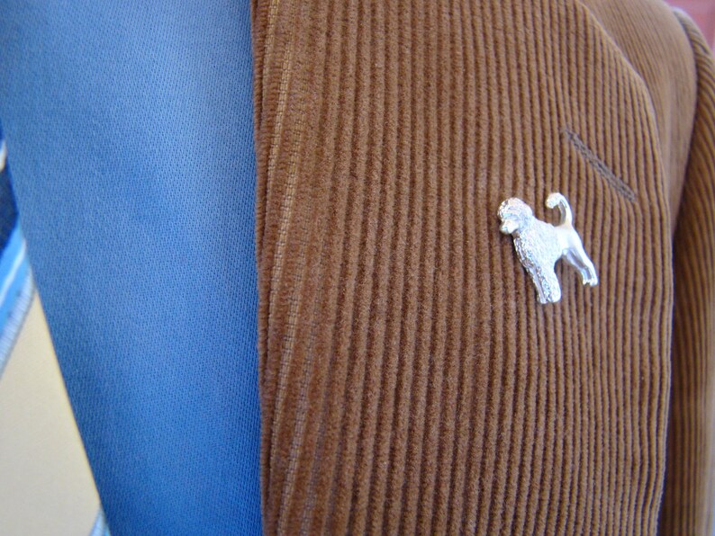 Puede incluir: Una chaqueta de pana marr&oacute;n con un broche de perro plateado en la solapa.