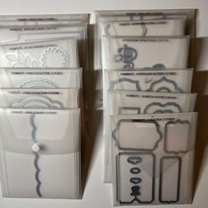 May include: A collection of clear plastic storage pouches, each containing metal die-cut shapes for crafting. The pouches are labeled with the names of the die sets, such as "Secret Garden" and "Labels Collection". The image shows a variety of shapes.