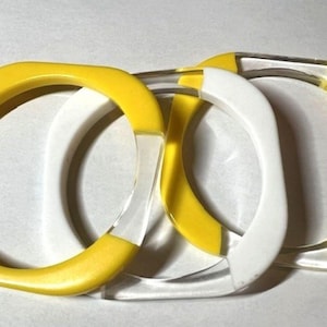 May include: Three square-shaped bangles. Two are yellow with clear, rectangular accents, and one is white with clear accents. The bracelets are made of a glossy material and are stacked together.