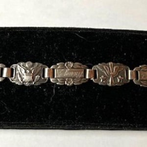 May include: Silver-toned link bracelet with rectangular and oval segments. The rectangular segments have engraved text, while the oval segments feature floral and heraldic designs. The bracelet is displayed on a black velvet surface.