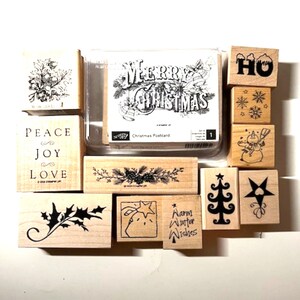 May include: A collection of wooden stamps with various Christmas-themed designs. The stamps include text like "Merry Christmas", "Peace, Joy, Love", and "Warm Winter Wishes", along with images of a Christmas tree, snowflakes, and a snowman.