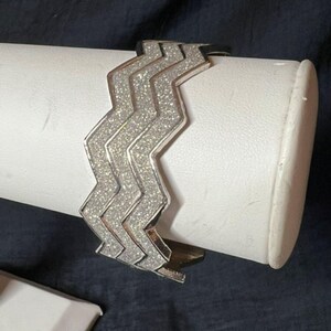 May include: A silver-toned cuff bracelet featuring a prominent zigzag or chevron pattern. The surface of the bracelet is covered in a fine, sparkling glitter texture that catches the light, creating a shimmering effect across the geometric design.