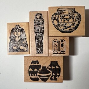 May include: A collection of wooden stamps featuring Egyptian-themed designs. The stamps include images of a pharaoh, a sarcophagus, hieroglyphs, and decorative vases. The designs are in black ink on rectangular wooden blocks.