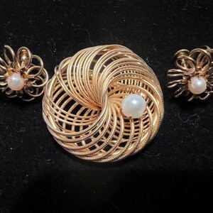 May include: A gold-toned jewelry set featuring a brooch and matching earrings. The brooch has a spiral design with a central pearl, and the earrings are floral-shaped with pearl centers. The set is displayed on a black surface.