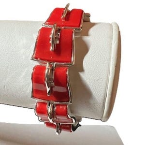 May include: A red and silver bracelet with a geometric design. The bracelet features rectangular red enamel links with silver-toned metal accents. The links are connected by silver-toned metal hinges. The bracelet is displayed on a white surface.