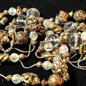 May include: Close-up of a necklace with a mix of clear, faceted beads and ornate, bronze-colored beads. The beads are strung on a delicate gold chain, creating a layered effect. The necklace has a vintage or antique aesthetic.