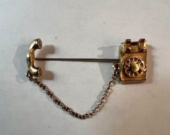 以下が含まれることがあります： 金色のヴィンテージ電話ピン。ピンは、チェーンで電話の受話器に接続されたロータリーダイヤル電話が特徴です。ピンはストレートピンに取り付けられています。