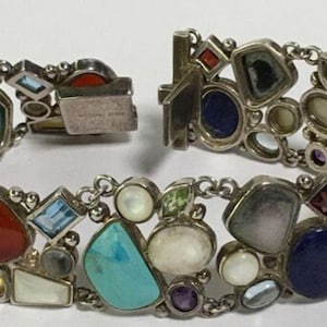 May include: Silver bracelet with a variety of colorful gemstones, including red, blue, turquoise, and white stones. The bracelet has a unique design with irregular shapes and a secure clasp.