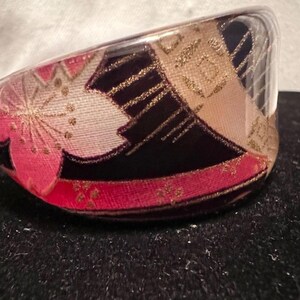 May include: A clear, curved bangle bracelet with an interior design featuring pink cherry blossoms, gold accents, and a red band. The bracelet has a black background and is likely made of resin or a similar material.