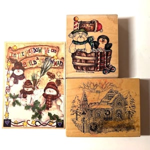 May include: Three wooden stamps with winter-themed illustrations. One features snowmen with the text "In the meadow we can build a snowman." Another shows a dog and penguin in a barrel. The third depicts a house.
