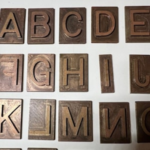 May include: A collection of vintage letterpress printing blocks. The blocks are made of a dark brown metal and feature the uppercase letters A through O. Each letter is raised and centered on a rectangular block.