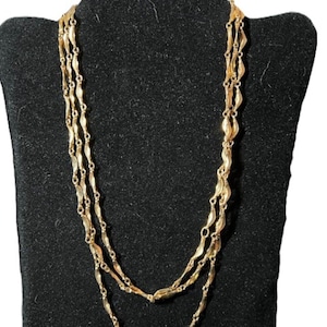 May include: A gold-toned necklace with three strands, each composed of small, elongated, leaf-shaped links. The necklace is displayed on a black jewelry bust, showcasing its layered design and intricate details. The necklace is a vintage piece.