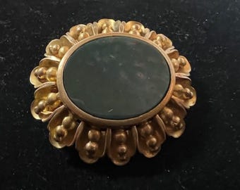 May include: A gold-toned brooch with a dark green oval stone set in the center. The brooch is surrounded by a decorative floral design.