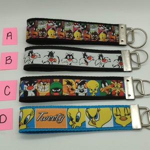 May include: Four keychains with cartoon character designs. Keychain A features a colorful pattern of various characters. Keychain B has a white background with Sylvester the Cat. Keychain C has a black background with various characters. Keychain D has a blue background with Tweety Bird and the word "Tweety".