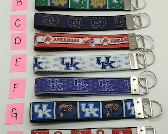 May include: Eight different keychains with various college logos. The keychains are made of a woven fabric with a variety of colors and designs. The logos include the University of Virginia Cavaliers, Notre Dame Fighting Irish, Arkansas Razorbacks, University of Kentucky Wildcats, and the University of Oklahoma Sooners.