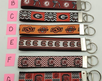 May include: Seven different keychains with various college logos. The keychains are made of black and white ribbon with the college logo printed on them. The keychains are attached to silver key rings.
