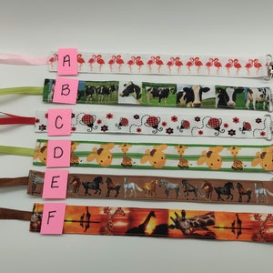 May include: Six different fabric baby teethers with colorful patterns and clips. The patterns include flamingos, cows, ladybugs, giraffes, horses, and giraffes in a sunset scene. The teethers are labeled A through F.