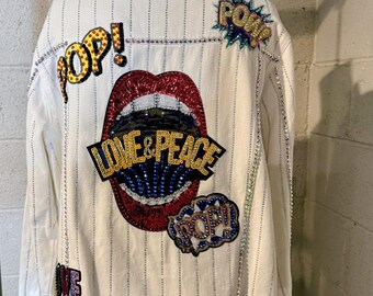 Op de afbeelding: Witte spijkerjas met verticale krijtstrepen en paillettenversieringen. Voorzien van patches met de woorden "POP!" en "LOVE & PEACE" in een kleurrijk paillettenontwerp. De jas heeft een gerafelde zoom.