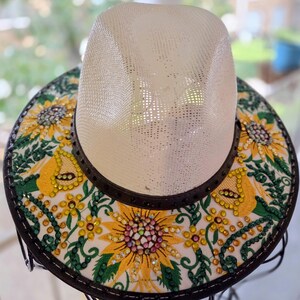 Peut inclure: Chapeau de paille avec une couronne blanche et un large bord orné de tournesols brodés. Le bord présente des motifs floraux jaunes, verts et roses avec des embellissements scintillants. Le chapeau a une bande en cuir marron foncé.