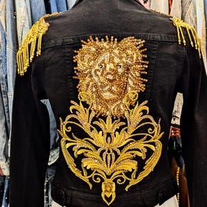 May include: Black denim jacket with gold embellishments. The back features a detailed lion design and ornate floral patterns, all in gold. The shoulders have gold fringe accents. A unique and stylish piece.