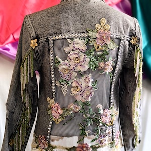 May include: A gray denim jacket with floral embroidery and beaded fringe. The embroidery features pink, purple, and green flowers with rhinestones. The fringe is made of green beads.