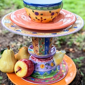 May include: A whimsical, tiered ceramic display featuring a teacup, saucer, and plates in vibrant colors. The design includes floral and geometric patterns, with a small teapot and decorative fruit accents.