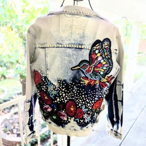 Peut inclure: Une veste en jean délavé clair avec un motif délavé à l'acide foncé, ornée au dos d'une grande application perlée représentant un colibri et des fleurs. Le design est agrémenté de nombreux strass transparents le long des coutures et du col.