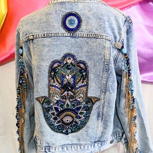 May include: A light blue denim jacket with a large, colourful hamsa hand design on the back. The hamsa is embellished with rhinestones, beads, and sequins. The jacket also features a smaller evil eye design on the back and gold chains with blue evil eye beads on the sleeves.