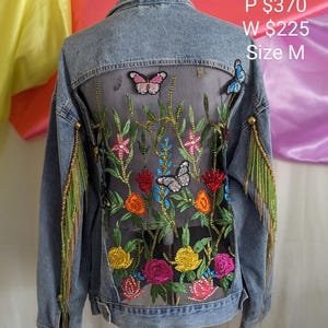 May include: A blue denim jacket featuring a back panel of sheer black striped fabric. The back is decorated with colourful beaded floral and butterfly appliqués. Long green and gold beaded fringe hangs from the sleeves. Text reads EK 262.
