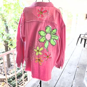 Könnte beinhalten: Eine leuchtend pinke Jeansjacke mit ausgefranstem Saum und Kragen. Die Rückseite ist mit vier großen Blumenapplikationen in Neongrün, Gelb, Orange und Pink verziert, die mit verstreuten klaren Strasssteinen und metallischen Verzierungen versehen sind.