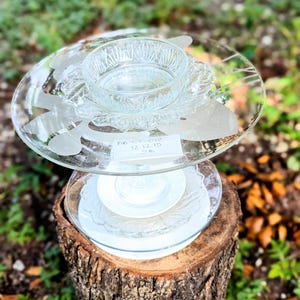 May include: A tiered glass serving dish, composed of clear glass plates and bowls, sits atop a tree stump. The top plate has etched designs. The arrangement is outdoors, surrounded by greenery and leaves. Text on a small tag reads "BB CY 012 12.12.15".