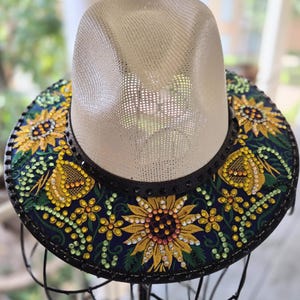 Peut inclure: Un chapeau de style fedora de couleur paille avec un bord décoratif. Le bord est bleu marine avec des broderies de tournesols jaunes, de feuilles vertes et de petits ornements scintillants. Le chapeau est présenté sur un support en métal noir.