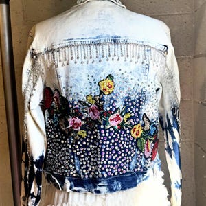 Peut inclure: Veste en jean bleu clair délavé, ornée de strass et de broderies florales. Le dos présente une cascade de strass et un motif floral vibrant avec des fleurs roses, jaunes et rouges. Les manches ont un effet tie-dye.