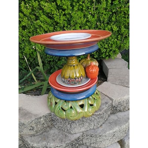 May include: A multi-tiered ceramic bird feeder with a green base, blue and red accents, and a white ceramic bird bath on top. The feeder is decorated with a green and yellow ceramic acorn and a small ceramic bird.