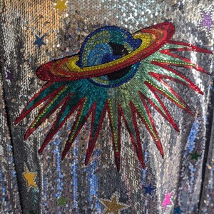 May include: A silver sequined jacket with a colorful embroidered design of a planet with rings and a starburst. The jacket is covered in silver sequins and has a variety of colorful stars scattered across the fabric.