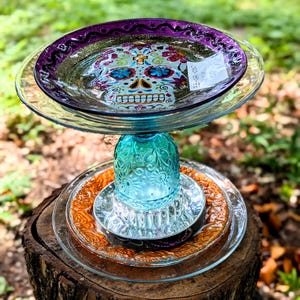 May include: A decorative bird bath with a Day of the Dead sugar skull design. The bird bath is made of glass and sits atop a tree stump. The top dish has a purple rim and the skull is decorated with colorful flowers.